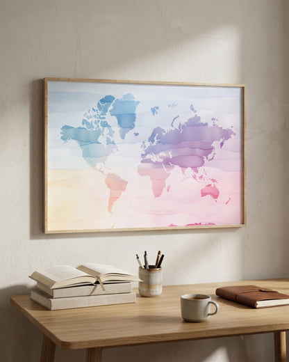 A framed world map watercolor print with pastel blue, purple, and pink tones hanging on a textured beige wall above a wooden desk with books, a mug, and a notebook.