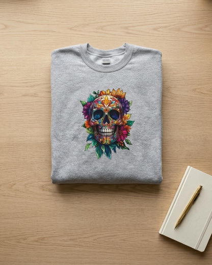 A folded light gray crewneck sweatshirt with a colorful sugar skull graphic on the front, displayed on a light wood tabletop next to a notebook and pen.