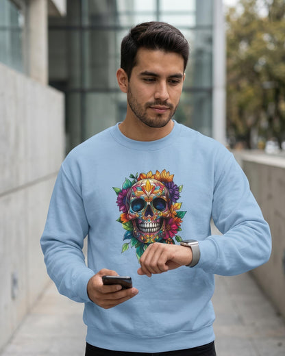 A man wearing a light blue crewneck sweatshirt with a colorful sugar skull and floral graphic on the front, standing outdoors in a modern urban setting.
