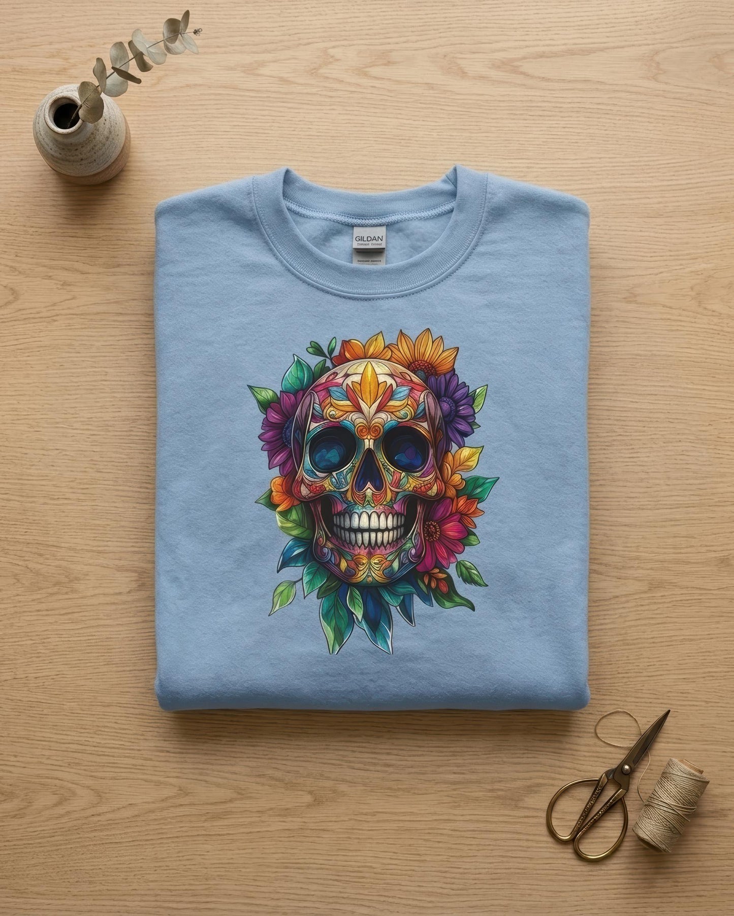 A folded light blue t-shirt with a colorful sugar skull graphic, displayed on a light wood background with decorative props including a small vase, eucalyptus sprig, scissors, and thread.