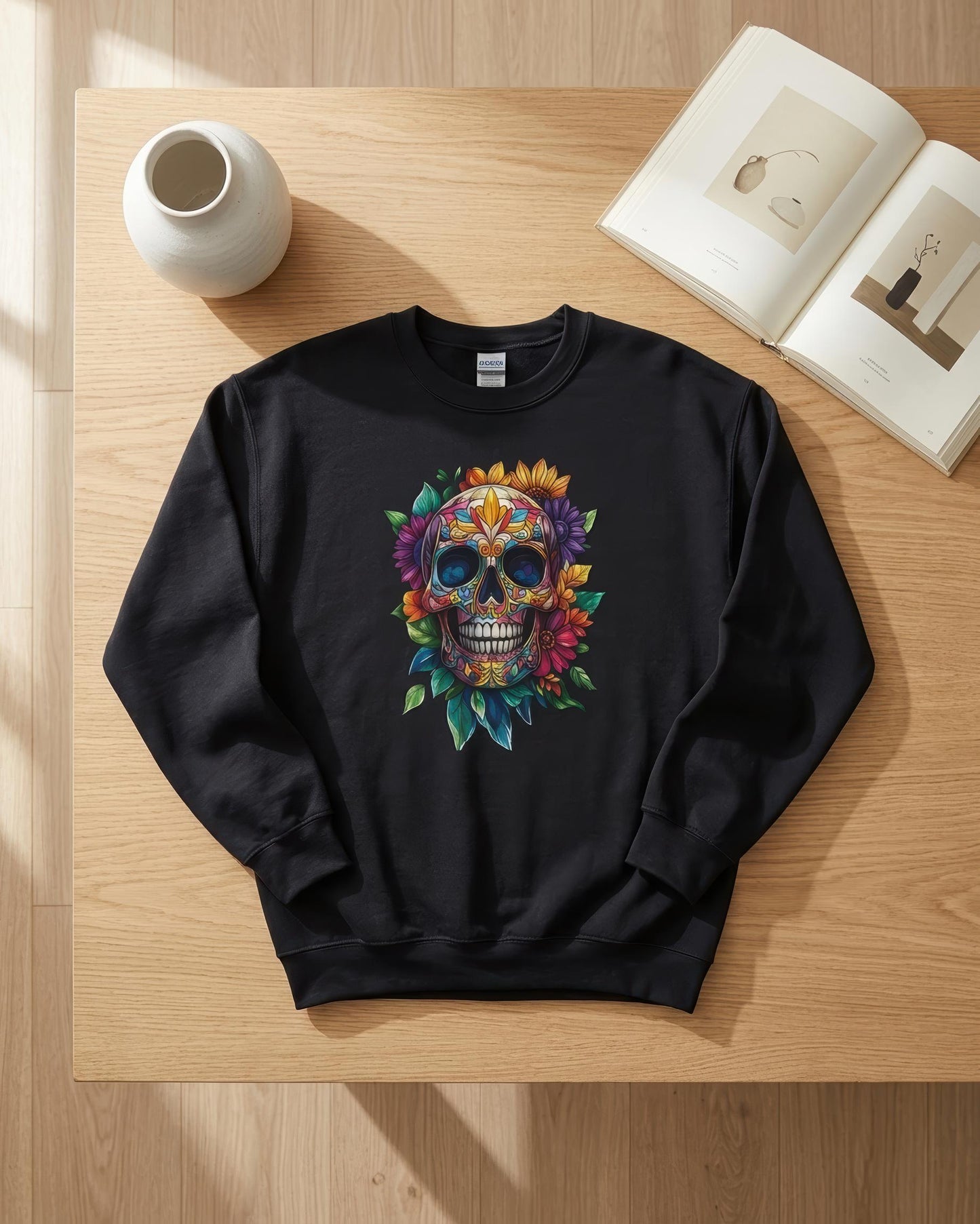 A black crewneck sweatshirt laid flat on a light wood table, featuring a colorful sugar skull graphic with flowers and leaves. The scene is styled with an open book, a small white vase, and natural sunlight casting soft shadows.
