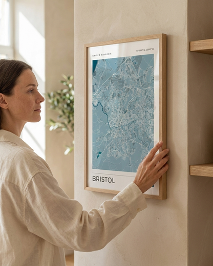 A woman in a cream linen shirt holds a framed city map print of Bristol against a warm beige wall in a modern interior setting.