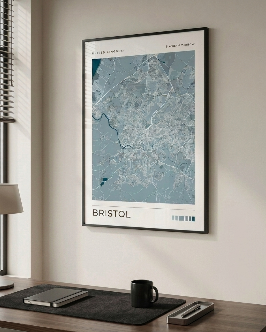 A framed city map print of Bristol, United Kingdom, displayed on a beige wall above a wooden desk with a laptop, notebook, and mug.