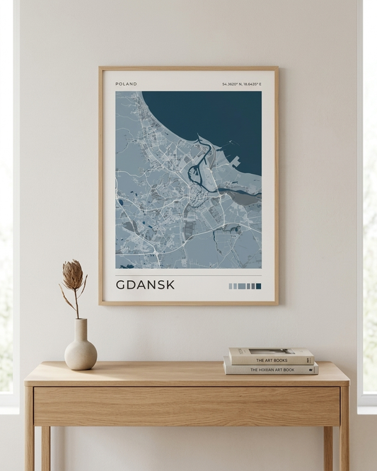 A framed city map print of Gdańsk, Poland, displayed on a light beige wall above a wooden console table with a vase and books.
