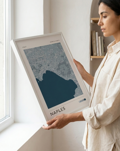 A woman in a cream linen shirt holds a large framed Naples city map print with a white border and dark blue water, standing by a bright window.