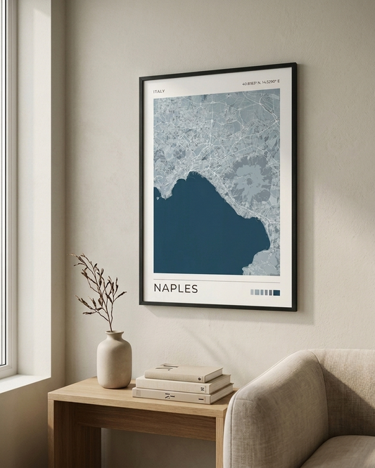 A framed Naples city map art print hangs on a beige wall above a wooden side table with a vase and books, next to a light-colored armchair.