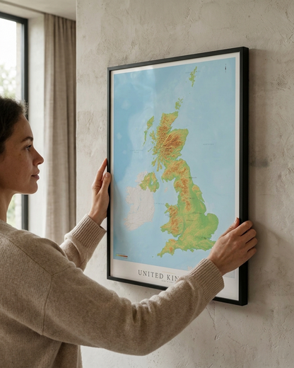 A woman in a beige sweater holds a framed topographical map of the United Kingdom on a textured light gray wall.