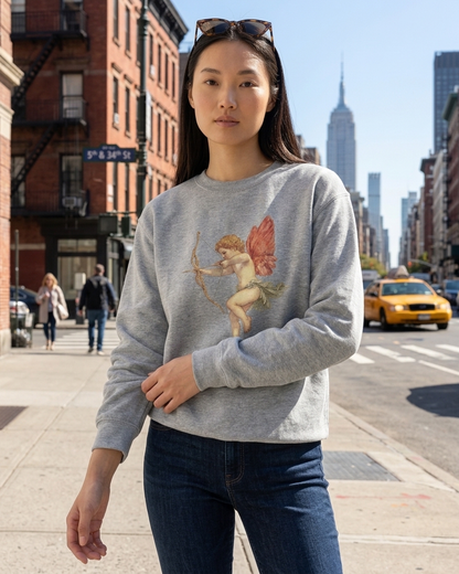 A woman wearing a light gray crewneck sweatshirt with a cherub graphic, standing on a city street with the Empire State Building in the background.