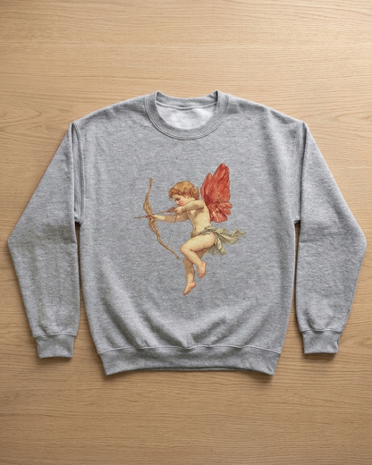 A heather gray crewneck sweatshirt featuring a vintage-style graphic of Cupid with red wings and a bow, laid flat on a light wood background.