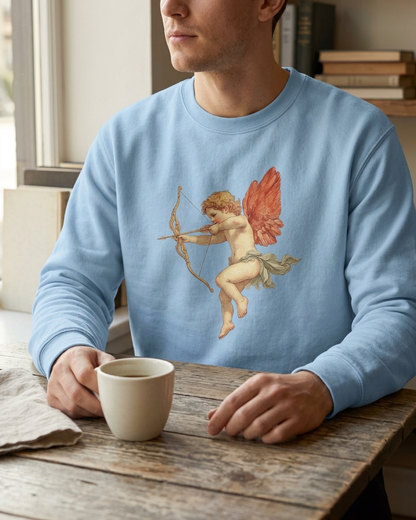 A man wearing a light blue crewneck sweatshirt with a graphic of Cupid, seated at a rustic wooden table holding a white mug.