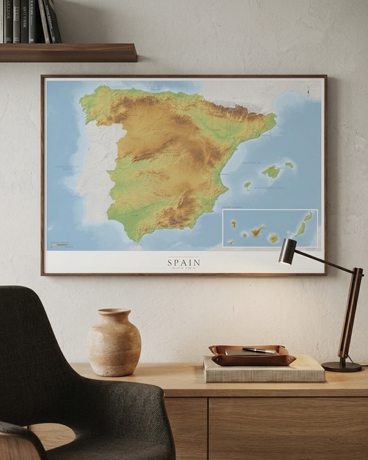 A framed topographic map of Spain and Portugal hanging on a light beige wall above a wooden desk with a chair, vase, and lamp.