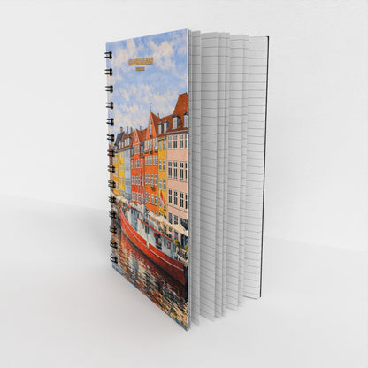 A spiral-bound notebook standing upright on a white surface, showing a colorful city canal scene with buildings and boats on the cover. The notebook has white lined pages visible on the right side.