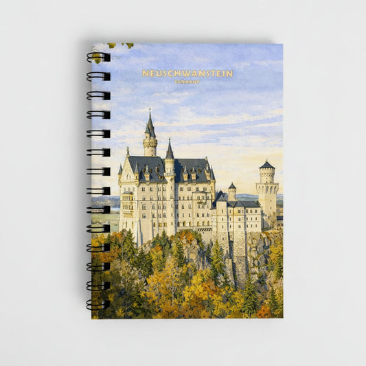 A spiral-bound notebook featuring a watercolor-style illustration of Neuschwanstein Castle in Germany, with the text 'NEUICHWANSTEIN GERMANY' printed at the top. The cover shows the castle above autumn-colored trees, and the notebook has black wire binding along the left side.
